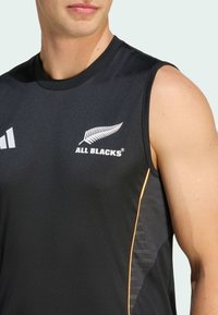 Svart ärmlös träningströja med "ALL BLACKS"-logotyp och ormbunksikon. Har orange detalj på sidan och strukturerat mönster. Tillverkad av mjukt tyg.