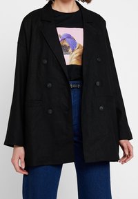 Černý lněný dvouřadý blazer s velkými knoflíky, klopovým límcem a bočními kapsami, vrstvený přes grafické tričko a džíny s vysokým pasem.