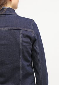 Czarna dżinsowa kurtka z wyraźnym kołnierzem, podwójnym przeszyciem i lekko dopasowanym krojem, o gładkiej fakturze i bez widocznych ozdób.