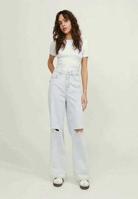 Light blue high-waisted jeans with distressed knee rips, paired with a fitted white short-sleeve top. Model stands against a neutral background.