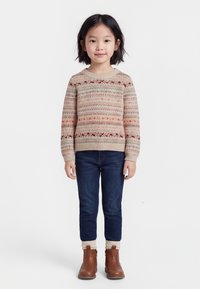 Noor tüdruk lühikeste tumedate juustega, kellel on beige mustriga kampsun, tumedad teksad, valged sokid ja pruunid pahkluu saapad, seisab valge tausta ees.