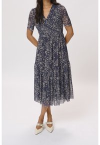 Marineblaue Blümchenkleid mit Wickeldesign, kurzen transparenten Ärmeln und gestuftem Rock. Getragen mit weißen spitzen Schuhen mit Riemenakzenten.