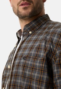 Gray and orange checkered button-up shirt with a collar, featuring white buttons and a chest pocket. Soft fabric texture.