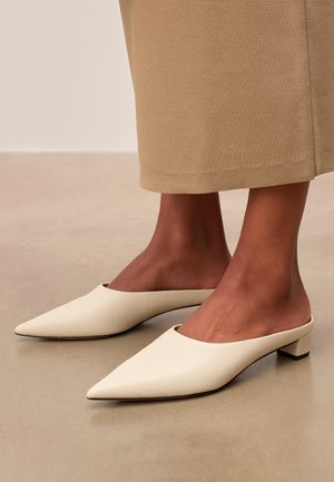 Moteris dėvi smėlio spalvos smailiais galais, žemo kulno mulei, derintus su tekstūruota smėlio spalvos suknele, stovinčią ant lygaus grindinio.