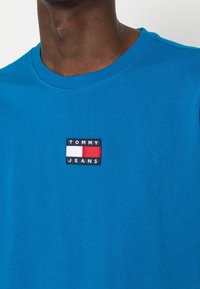 Niebieska bawełniana koszulka z okrągłym dekoltem oraz naszywką z logo "TOMMY JEANS" w czarnym, białym i czerwonym kolorze.