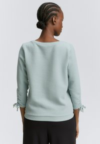 Svetlomodrý textúrovaný sveter s tromi štvrtinovými rukávmi, s nastaviteľnými viazaniami na manžetách a uvoľneným výstrihom. Nosí sa s čiernymi nohavicami.