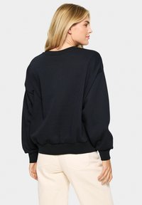 Čierny oversized sveter s rovnými okrajmi, so spustenými ramenami a rebrovanými manžetami a lemom. Nosený s svetlo sfarbenými nohavicami rovného strihu.
