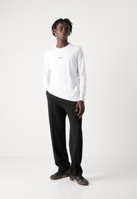 Camisa branca de mangas longas com um pequeno logótipo preto, combinada com calças pretas e sapatilhas escuras. Design minimalista, textura suave.