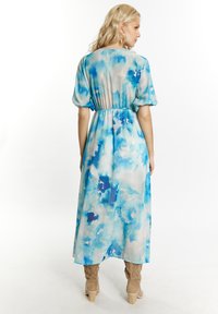 Abito maxi in tessuto leggero e trasparente con stampa ad acquerello blu. Presenta maniche a palloncino e vita elasticizzata. Indossato con stivali color beige al ginocchio.
