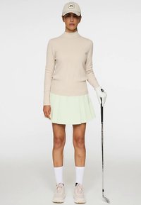 Beiger, gerippter Pullover mit langen Ärmeln, hellgrüner plissierter Rock, weiße knielange Socken, beige Turnschuhe und eine beige Kappe mit Logo. Golfclub in der Hand.