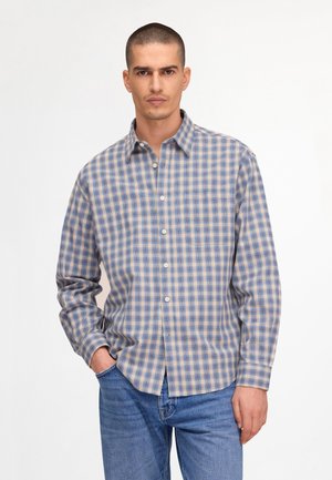 Mann trägt ein blaues, beiges und weiß kariertes Hemd mit Knöpfen und blaue Jeans, steht mit einer Hand in der Tasche vor weißem Hintergrund.