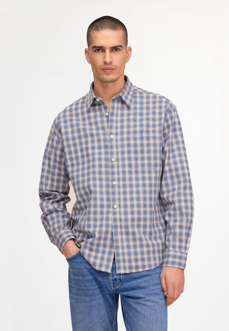 Homme portant une chemise à carreaux bleue, beige et blanche à boutons et un jean bleu, debout avec une main dans la poche sur un fond blanc.