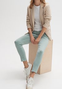 Femme assise sur un cube en bois portant un blazer beige, une chemise blanche, un pantalon vert clair et des baskets blanches avec des lacets.