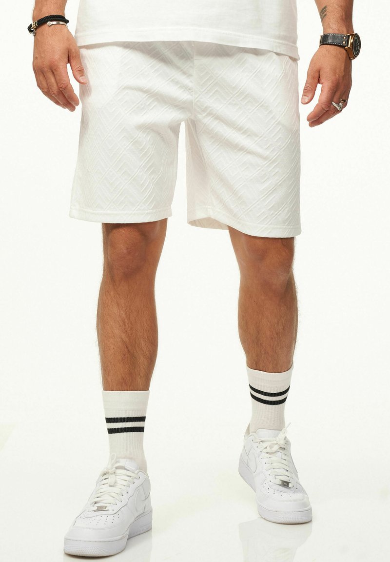 Weiße, gemusterte Shorts mit einem strukturierten Diamantdesign, geradem Schnitt und dezenter Saum, kombiniert mit weißen Sneakers und gestreiften Socken.