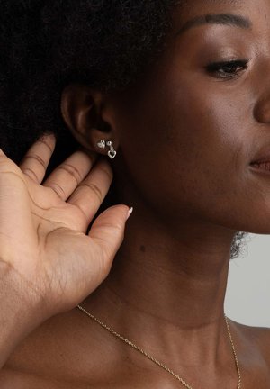 Silverörhängen i hjärtform, med en större och en mindre, som bärs av en mörkhyad person, tillsammans med ett fint guldhalsband.