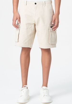 Un short cargo beige en coton, doté de deux poches latérales, d'une fermeture à bouton à l'avant et d'une coupe décontractée, associé à des baskets blanches.
