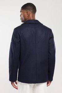 Granatowy wełniany płaszcz z klasycznym kołnierzem i długimi rękawami, z subtelną teksturą i jednym rozcięciem z tyłu, noszony z kremowymi spodniami.