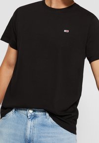 Musta puuvillapaita, jossa on lyhyet hihat ja pyöreä kaula-aukko, rintakehässä on pieni punavalkoinen brodeerattu logo.