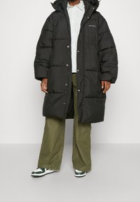 Zwarte pufferjas met capuchon, voorzien van een rits en drukknopen, gestepte design en zijzakken; gedragen over een gestreept shirt en groene broek.