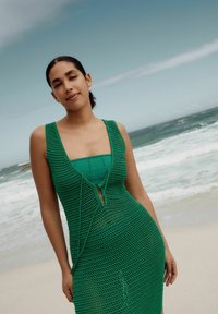 Groene gehaakte jurk met een v-hals en een strikdetail in de taille, gelaagd over een effen groene aansluitende jurk. Zachte gebreide textuur, aansluitende silhouette.