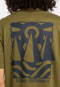 T-shirt vert olive avec un graphique bleu marine représentant trois montagnes triangulaires au-dessus de vagues courbées et le texte "ELEMENT SKATEBOARD CO." en bas.