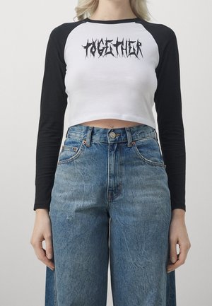 Vrouw die een zwart-wit crop top met lange mouwen en de tekst "TOGETHER" draagt, gecombineerd met een hoge taille spijkerbroek, staand tegen een effen achtergrond.