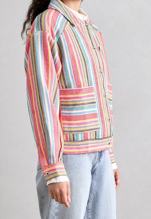 Person trägt mehrfarbige gestreifte Knopfleistenjacke mit großen aufgesetzten Taschen über einem weißen Hemd und hellblauen Jeans.