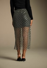 Schwarzer und weißer transparenter Rock mit einem Rautenmuster, knielang, mit einem Schlitz versehen, über einem taillierten Oberteil getragen und mit schwarzen Absätzen kombiniert.