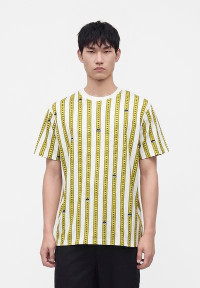 Homme portant une chemise blanche à manches courtes avec des rayures verticales jaunes en forme de ruban à mesurer, debout devant un fond clair uni.