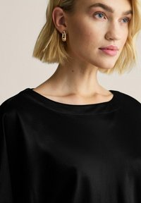 Sort satin top med bred udskæring, glat tekstur og draperet design, parret med en metallisk guldfarvet ørering, sat mod en neutral baggrund.
