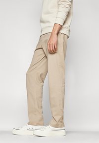 Pantaloni beige su misura con taglio dritto, realizzati in tessuto misto cotone. Indossati con sneakers bianche con dettagli neri.