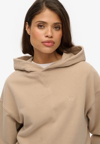 Sweat-shirt à capuche beige avec une coupe décontractée, doté d'une poche kangourou et d'un brodé logo subtil sur la poitrine gauche. Texture douce.
