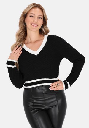 Vrouw glimlachend, gekleed in een zwarte V-hals trui met witte bies en zwarte leren broek, poserend met één hand op de heup en de andere die het haar raakt.
