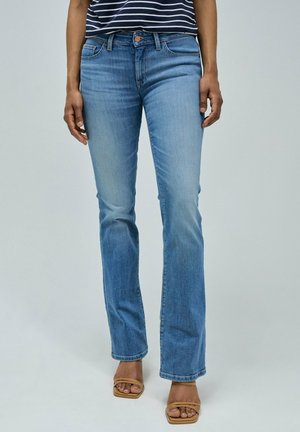 Jean en denim bleu clair évasé avec un lavage délavé, cinq poches et une fermeture à bouton en cuivre. Porté avec des sandales à talons avec des lanières marron.