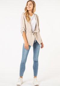 Beige lichtgewicht jack met een strik taille, opgerolde mouwen en een ontspannen pasvorm, gecombineerd met een witte t-shirt en lichtblauwe skinny jeans.