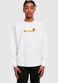 Hvid langærmet T-shirt med et farverigt grafik af en gravhund i en gul frakke og rød hat. Stoffet ser blødt og let ud.