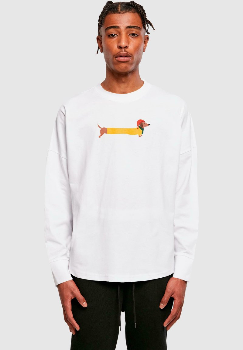 Camiseta blanca de manga larga con un gráfico colorido de un dachshund con un abrigo amarillo y un sombrero rojo. La tela parece suave y liviana.