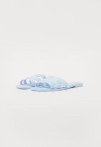 Ljusblå platta sandaler gjorda av mjukt material, med tre böjda remmar och en platt sula med en subtil logotypdetalj.