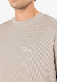 Hanorac bej cu nervuri, cu guler rotund, având un logo brodat alb. Materialul are un aspect texturat, moale.