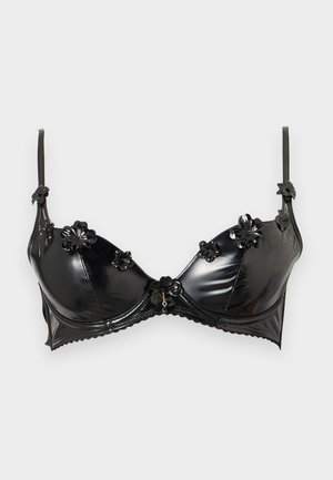 Kobieta stoi boso, ubrana w czarny satynowy biustonosz z kwiatowymi detalami i dopasowane majtki z koronkową lamówką na jednolitym białym tle.