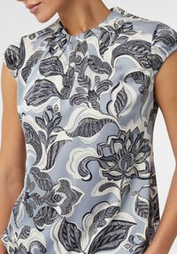 Kurzärmlige Bluse aus hellblauem Stoff mit aufwendigen schwarzen Blumen- und Blättermustern, versehen mit einem gerafften Halsauschnitt.