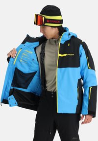 La giacca da sci blu e nera presenta accenti gialli, tasche con zip e un cappuccio. L'interno è dotato di dettagli stampati e una tasca in rete.