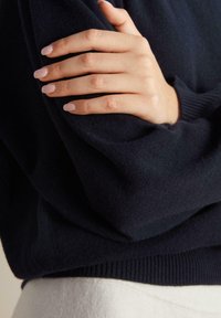 Marinblått stickat tröjtyg med ribbad mudd, delvis täckt av en hand med ljusrosa nagellack som vilar på ärmen.