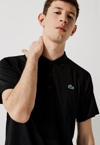 Černé polo tričko se třemi knoflíky, krátkými rukávy a zeleným vyšitým krokodýlem na levé straně hrudi. Hladká textilie.