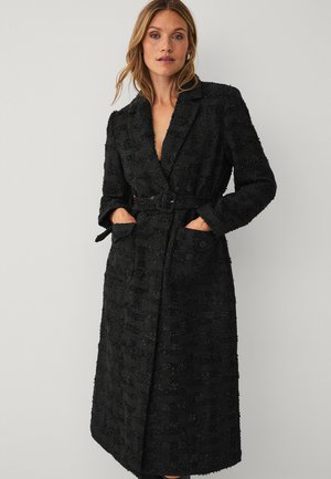 Kvinde iført en lang sort tekstureret frakke med bælte, revers, lommer og knappedetaljer, stående foran en ensartet lys baggrund.