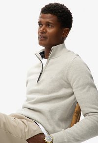 Mand med kort krøllet hår iført lys grå striktrøje med lynlås, beige bukser og guld armbåndsur, sidder på en træstol og kigger til siden.