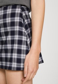 Námorně modrá a bílá kostkovaná sukně s rozšířeným tvarem, vyrobená z měkké látky. Má boční kapsy a lem nad středem stehna.