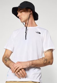 Weißes T-Shirt mit kurzen Ärmeln und schwarzem Logo auf der Brust, kombiniert mit einem schwarzen Bucket Hat, der ein verstellbares Kinnband und Belüftungsöffnungen hat.