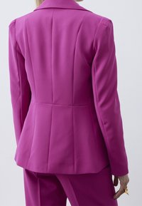 Blazer ajusté en tissu rose vif, avec épaules structurées, col à revers cranté et coutures arrière détaillées, accompagné d'un pantalon assorti.