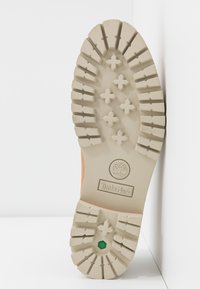 Semelle de botte Timberland beige avec de profondes rainures, des motifs croisés et un petit détail hexagonal vert près du talon.